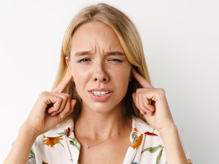 Unzufriedenes schönes Mädchen, das sich die Ohren zuhält, während es vor einem weißen Hintergrund isoliert vor der Kamera posiert.