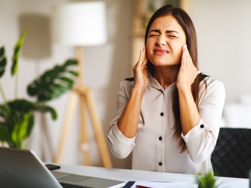Eine junge Frau sitzt an einem Schreibtisch im Büro und hat Kopfschmerzen, während sie mit einem Laptop arbeitet.